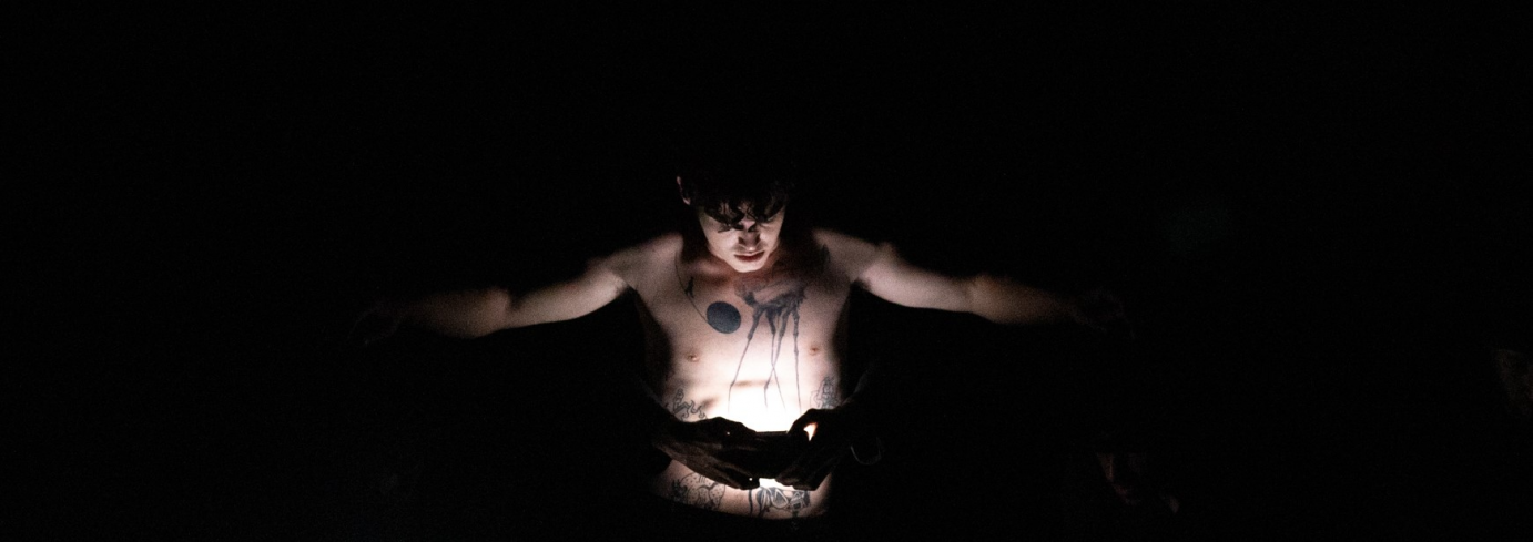 A man with tattoos stands, reaching his hands outward.